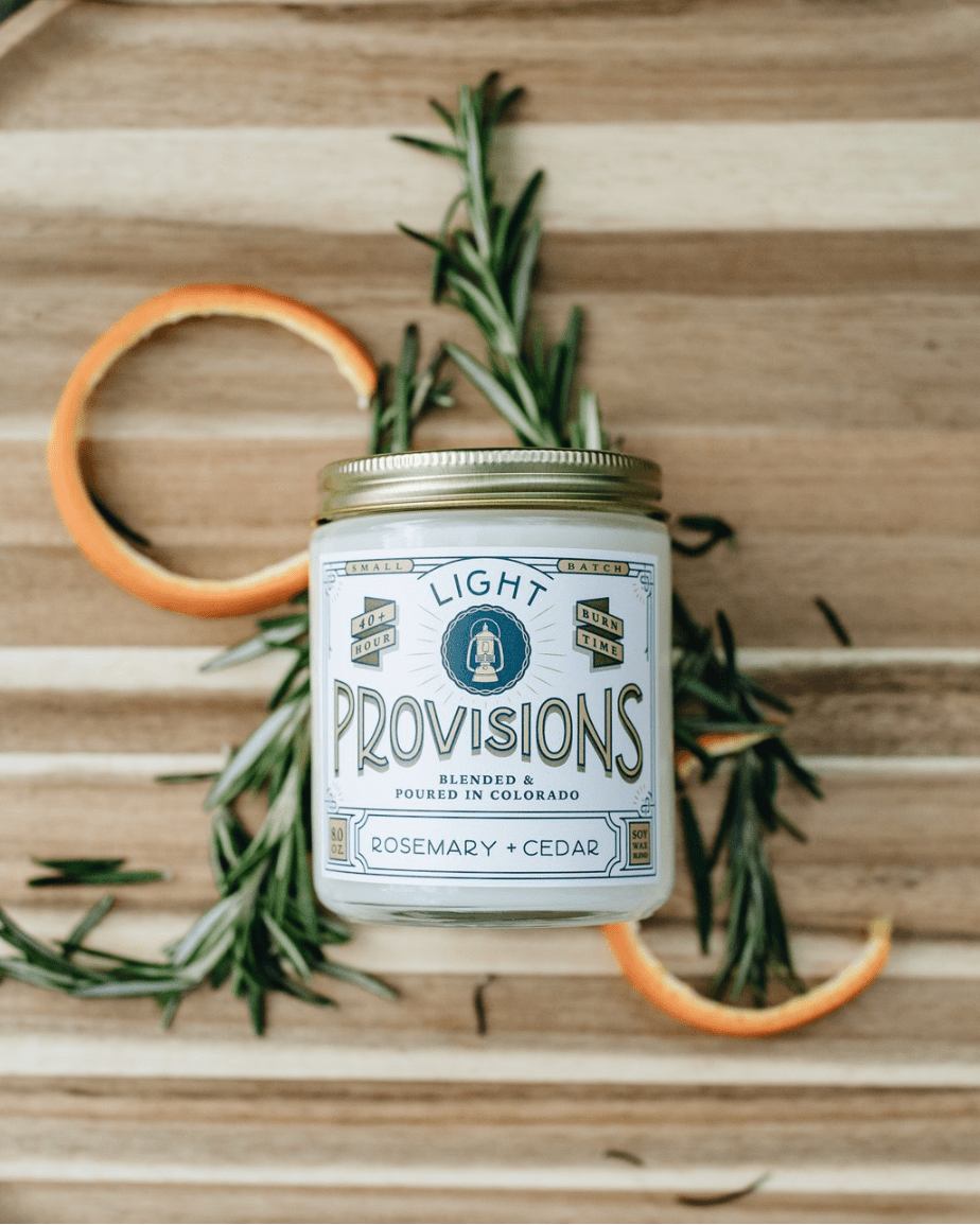A jar candle labeled 'Light Provisions' with rosemary and cedar scent, styled on a wooden surface with rosemary sprigs and orange peel slices.