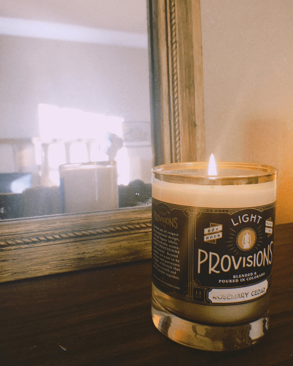 A lit candle in a clear glass jar with a dark label reading Light Provisions, placed on a wooden surface in front of a mirror.