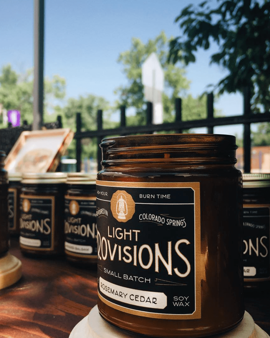 A jar of Colorado Springs Light Provisions Rosemary Cedar candles on a wooden table, with other matching jars blurred in the background outdoors.