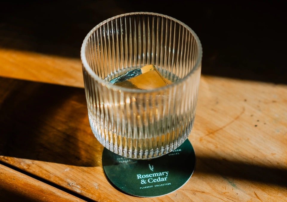 Ribbed glass tumbler with a clear cocktail and ice, photographed on a wooden surface resting on a Rosemary & Cedar dust cover