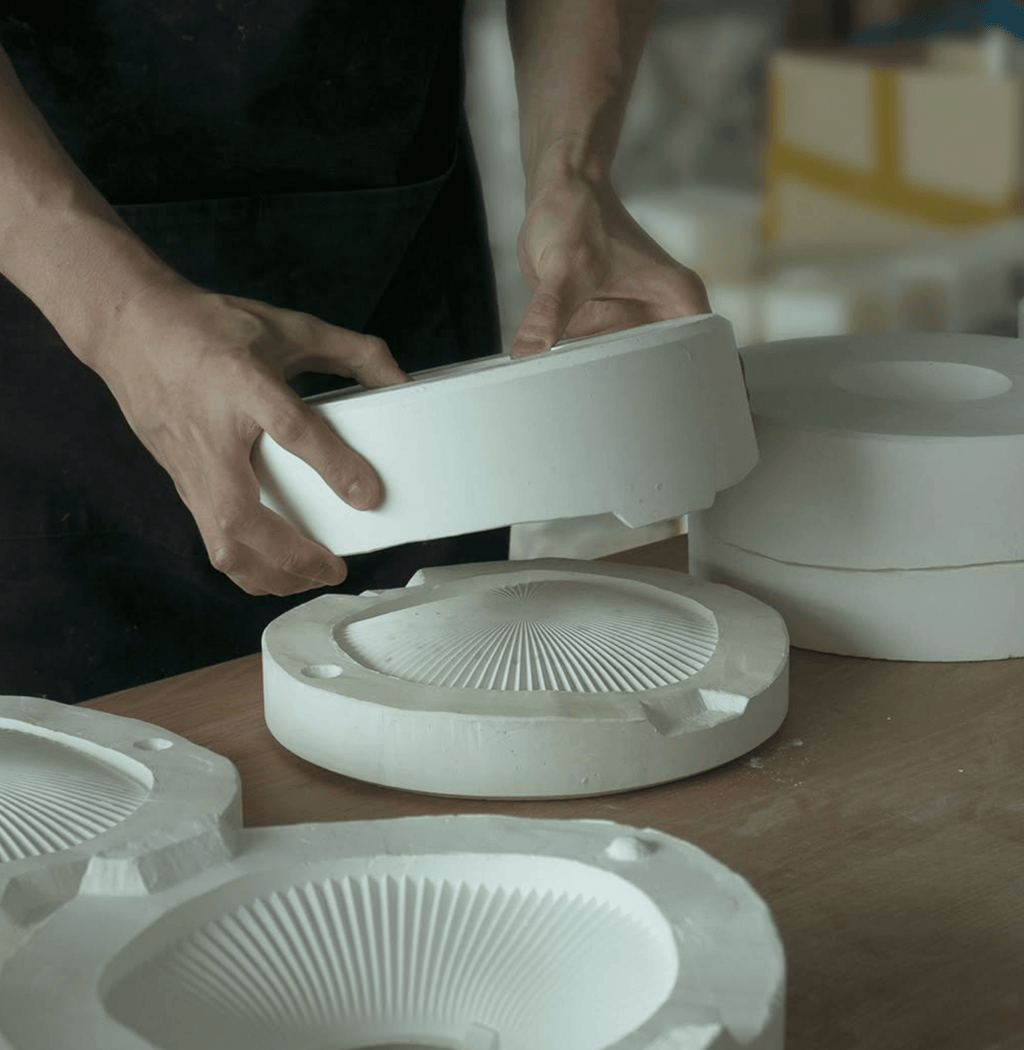 Hands assembling white ceramic incense holders with a mold on a worktable surrounded by other handmade components.