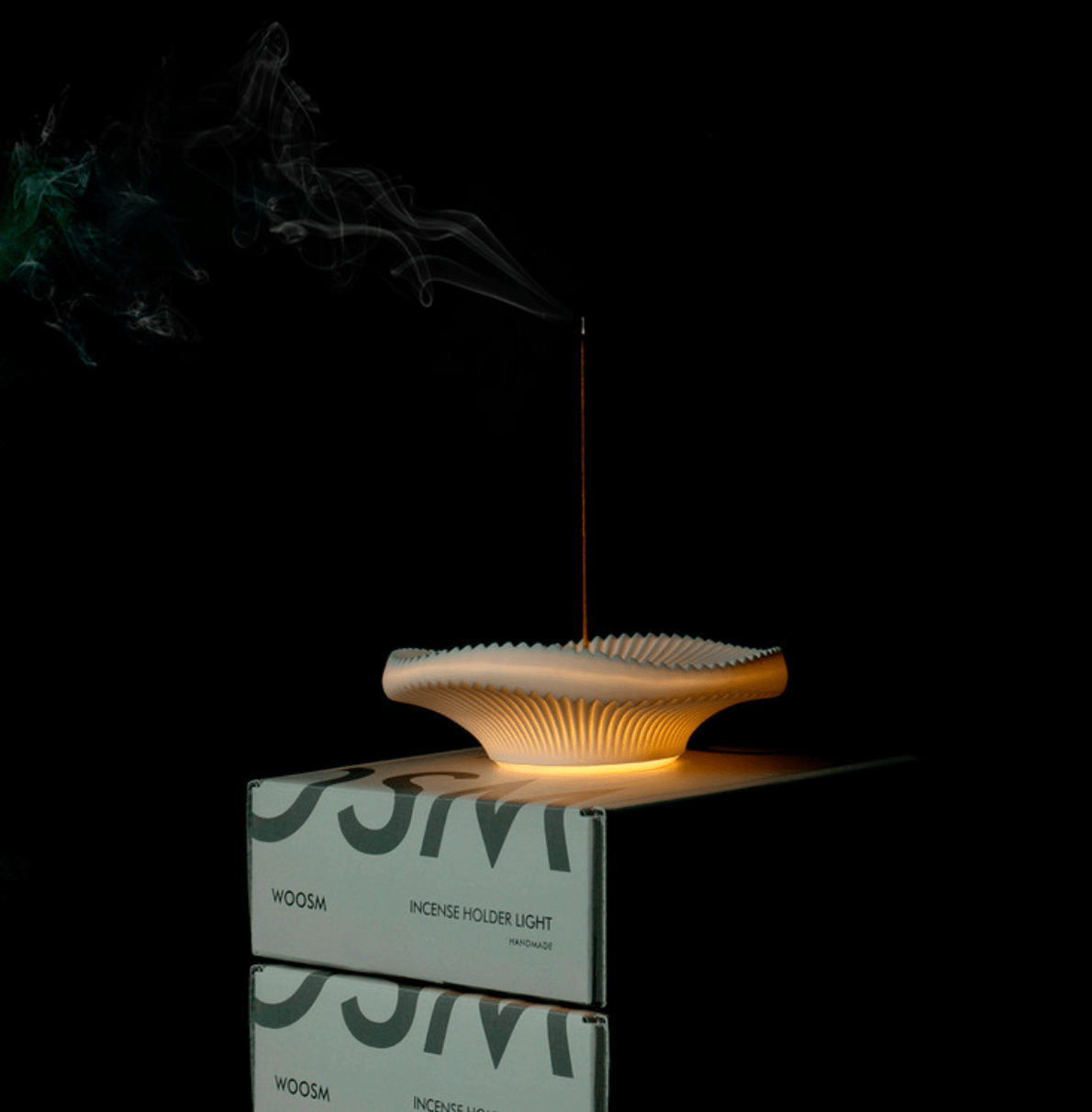Single incense stick burning in a glowing ceramic holder against a dark background. Holder is resting on Woosm gift box