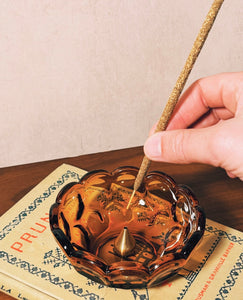 Hand placing an incense stick into a decorative glass bowl on a wooden surface.