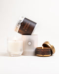 Tobacco Walnut glass tumbler candle beside matching travel tin on white background, both lit