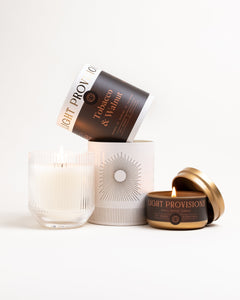 Tobacco Walnut glass tumbler candle beside matching travel tin on white background, both lit