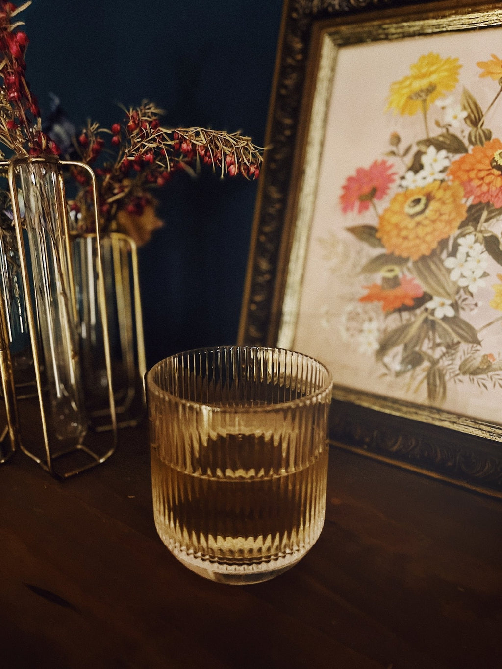 Ribbed clear glass with an amber drink on a wooden table, vintage decor and framed artwork softly blurred in the background.