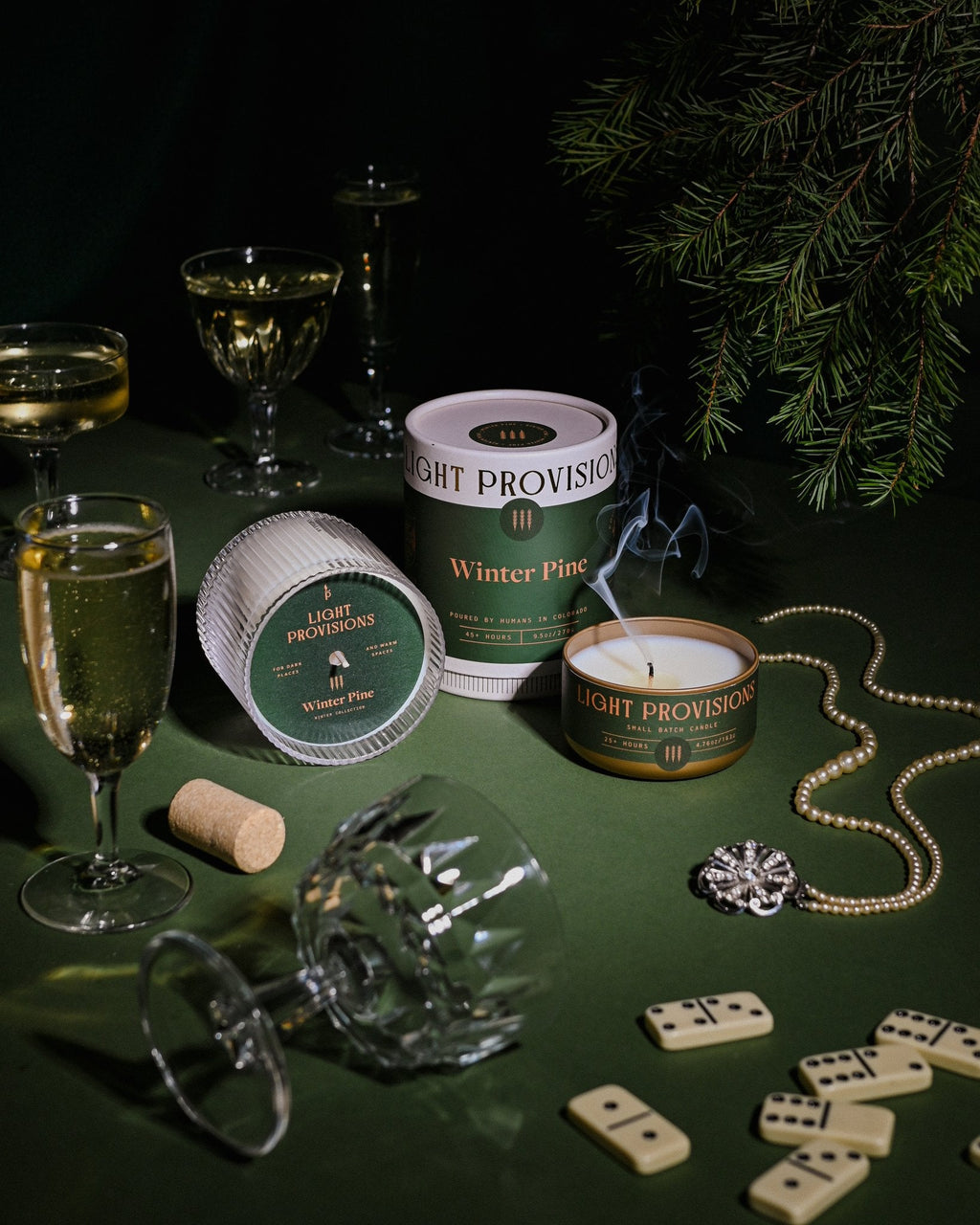 Winter Pine candle with packaging surrounded by champagne glasses, pine branches, dominoes, on dark green table.