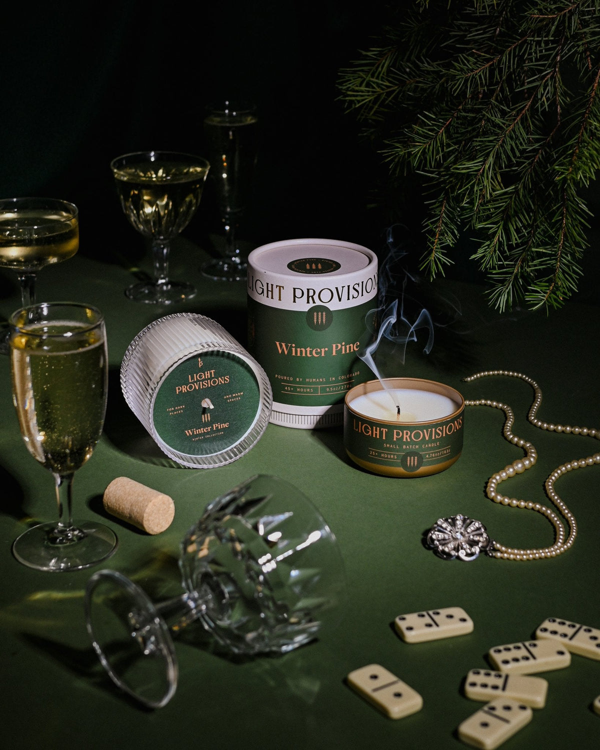 Winter Pine candle with packaging surrounded by champagne glasses, pine branches, dominoes, on dark green table.