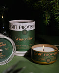 Winter Pine candle, box, & lit travel tin beneath pine greenery with champagne glass nearby
