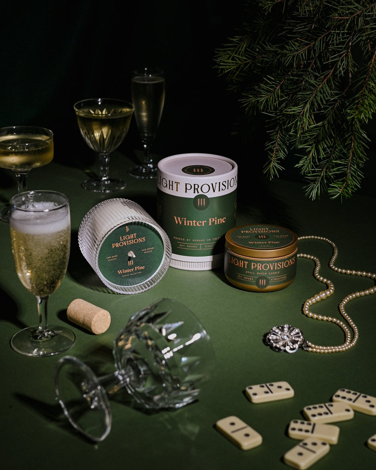 Winter Pine candle set with crystal goblets, dominoes, and pine branches, styled like a holiday evening table scene.