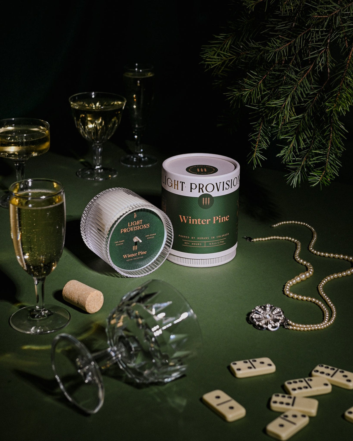 Winter Pine candle beside crystal glassware, cork, and string of pearls under evergreen branches