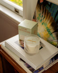 Grapefruit & Oakmoss candle in crystal glass resting on stacked books beside a sunlit window