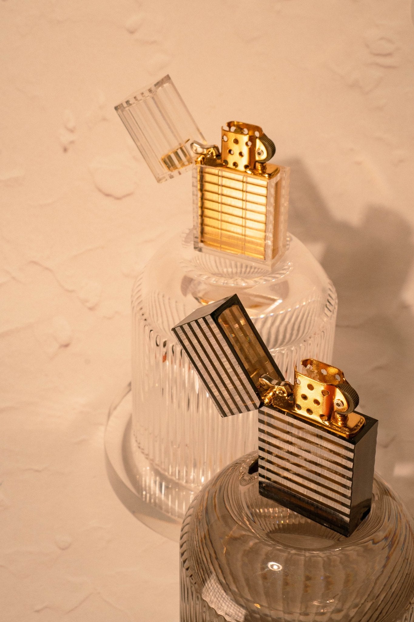 Two Latitude Hard-edge Lighters by Tsubota Pearl in clear and black with their gold parts visible, sitting on glassware