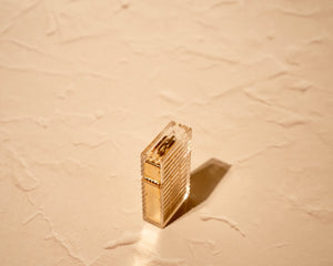 Clear Tsubota Pearl lighter casting striped shadow pattern under warm light, lid closed, brass elements exposed in warm light


