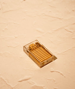 Tsubota Pearl Hard Edge lighter in clear ribbed polycarbonate casing, lid closed on textured reflective neutral surface. Brass parts visible under casing. Lighter laying on its side exposing bottoms screw, cotton wadding, and spare flint.