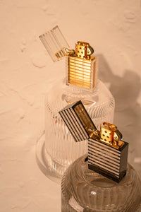 Latitude Lighters in both black and clear shown with their lids open, gold brass elements under clear poly casing, sitting on inverted rippled Light Provisions glassware.