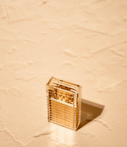 Tsubota Pearl Hard Edge lighter in clear ribbed polycarbonate casing, lid closed on textured reflective neutral surface. Brass parts visible under casing
