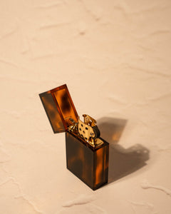 Tsubota Pearl Hard Edge lighter in amber casing, open lid, shown on textured beige surface