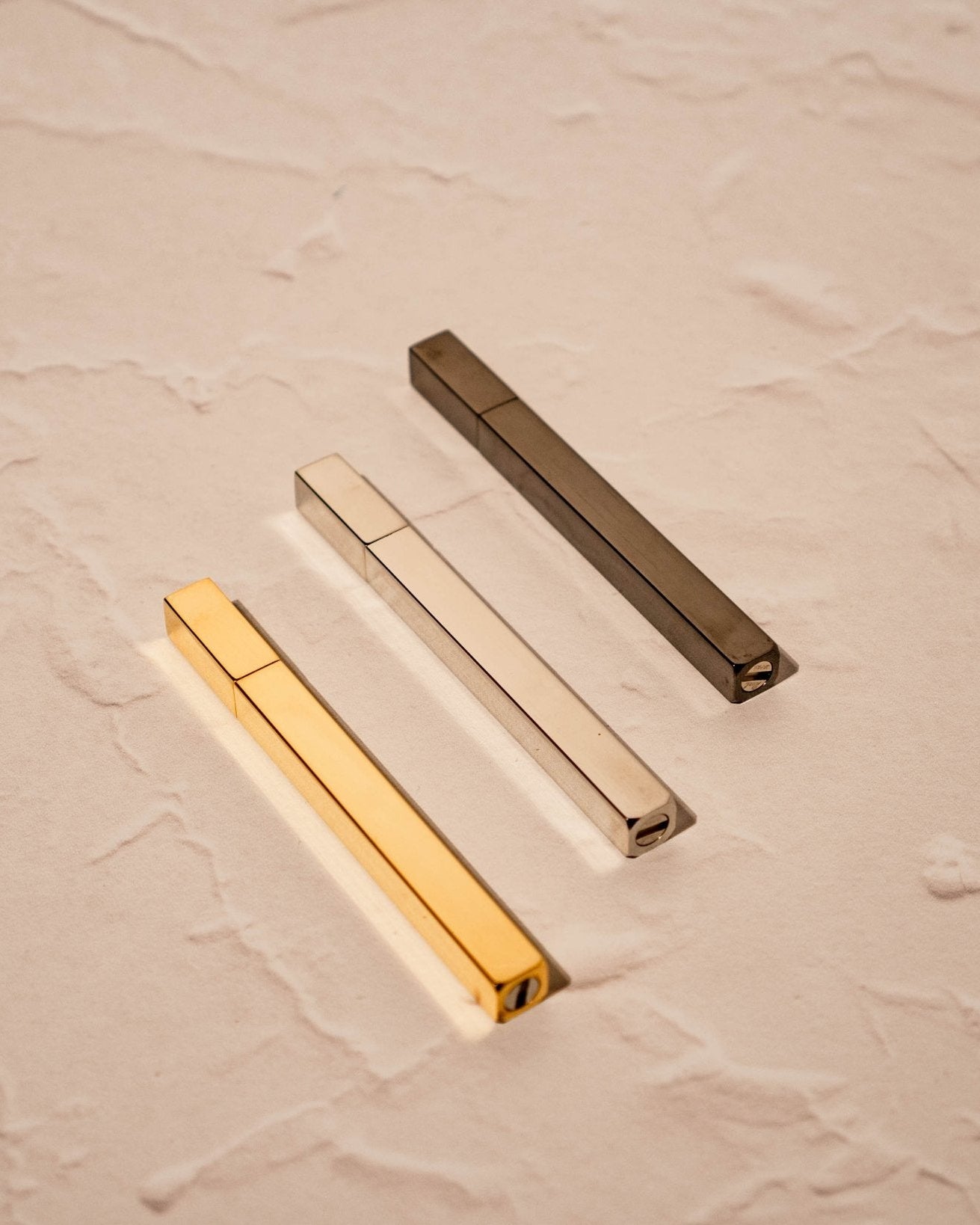 Three Queue Lighters by Tsubota Pearl in silver, gold, and brass finishes on beige surface