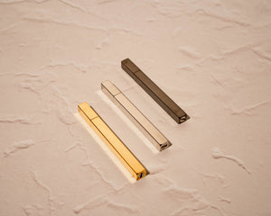 Array of 3 Queue stick lighter laying side by side - polished gold, silver, and black in view.