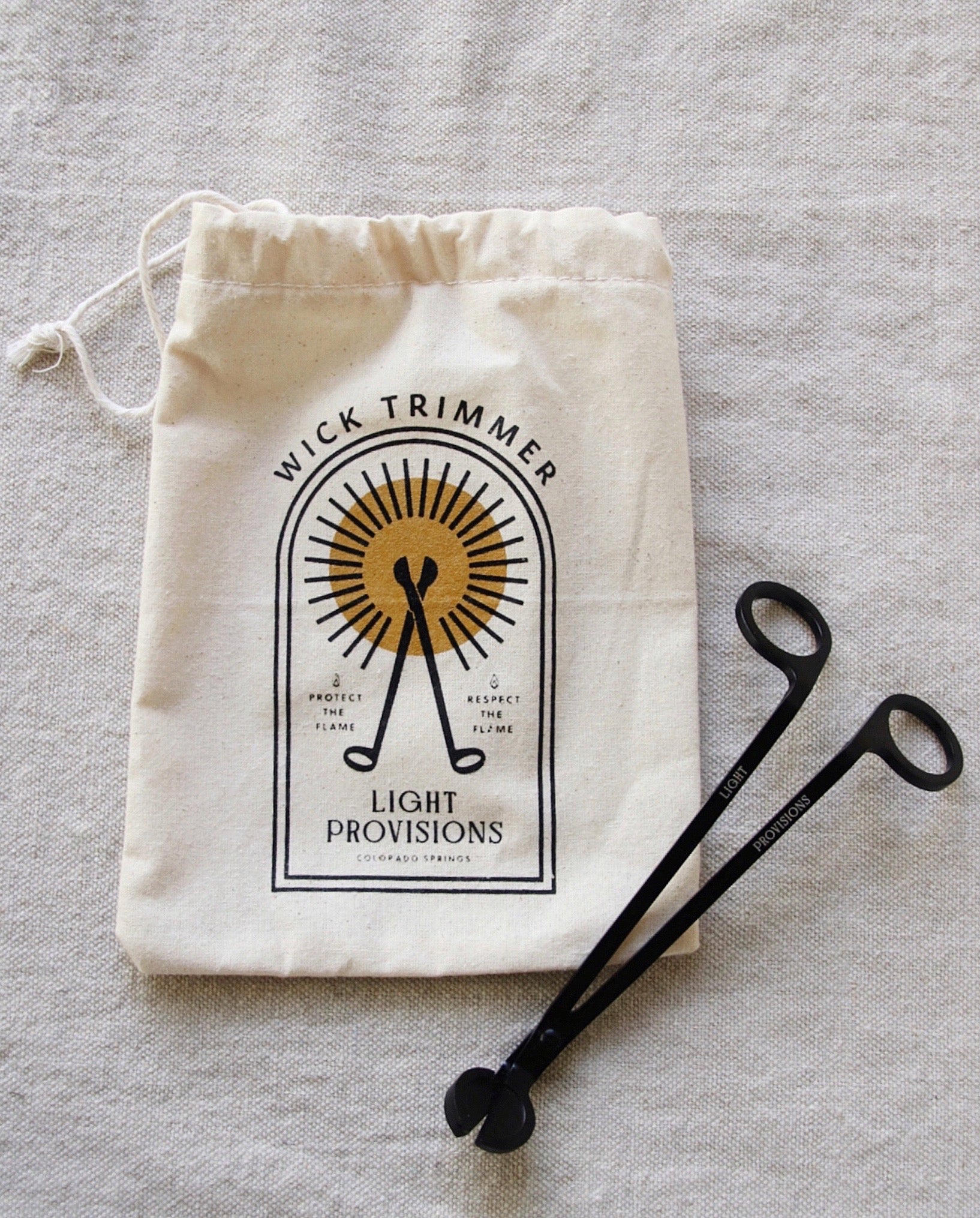 Black metal wick trimmer placed next to Light Provisions cotton storage bag with golden sunburst logo.