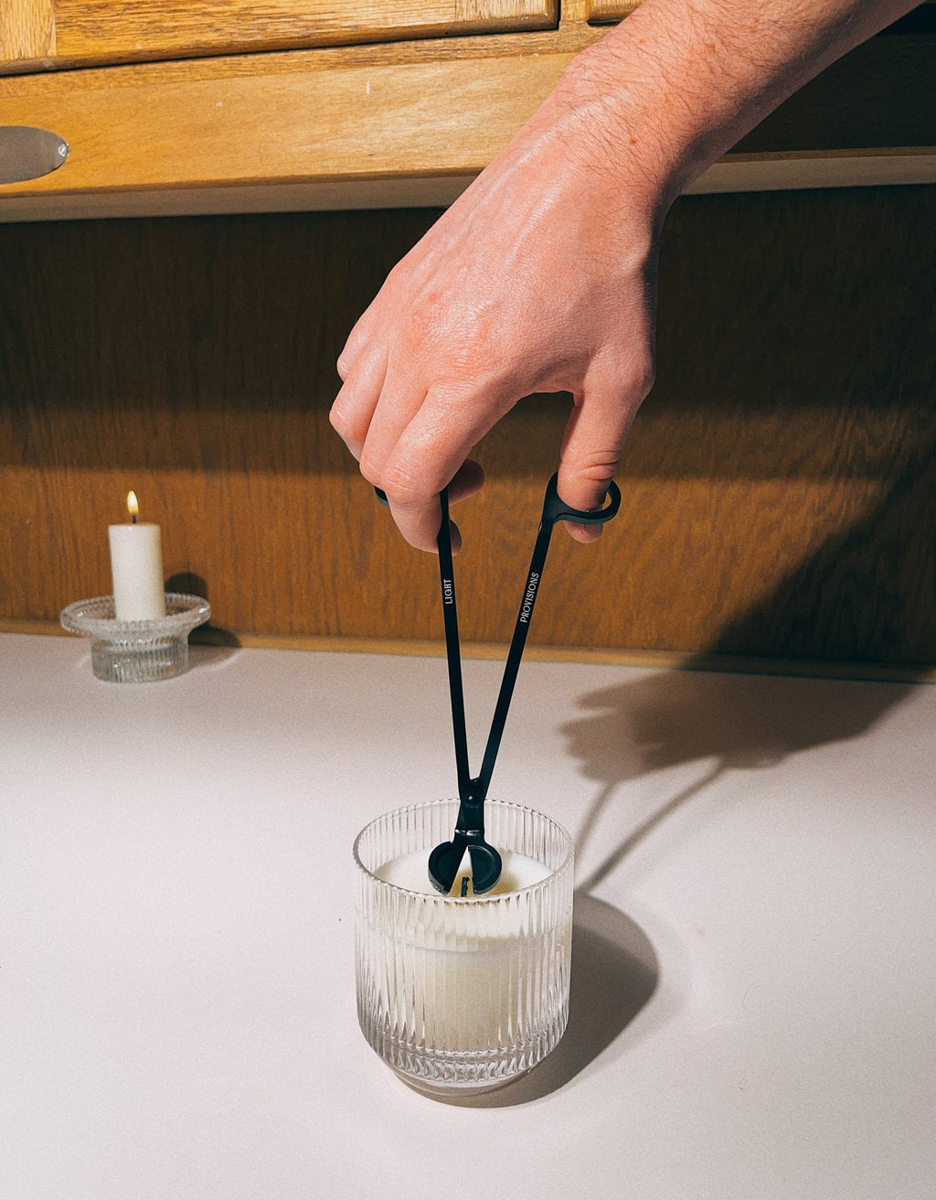 Person using black wick trimmer to trim candle wick in a white ribbed glass vessel on a wooden table.