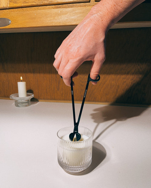 Person using black wick trimmer to trim candle wick in a white ribbed glass vessel on a wooden table.