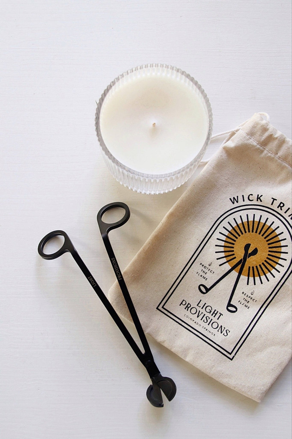 Overhead view of black wick trimmer, Light Provisions cotton storage bag, and white candle on a bright background.